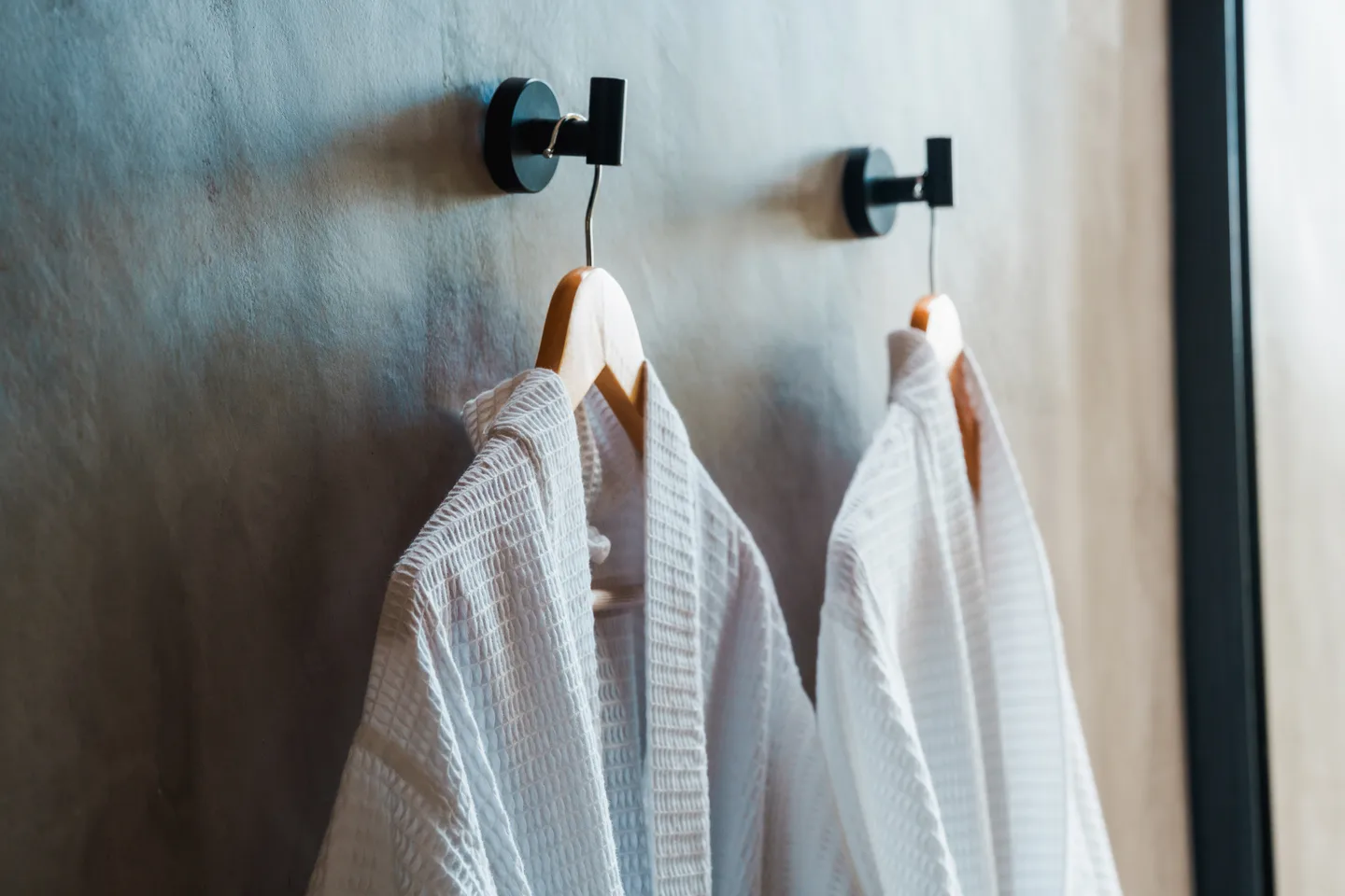 Two white waffle bathrobes on timber hangers against a polished concrete wall at NEST Los Cabos